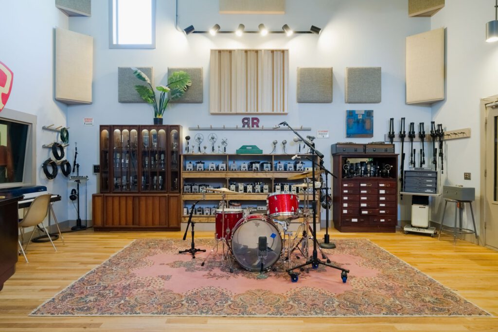 Recording studio live room with drum set on rug, vintage equipment, and sound treatment panels.
